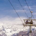 A snowy landscape in Auli, Uttarakhand, featuring ski slopes, a ropeway tower, and majestic snow-capped Himalayan peaks in the background.