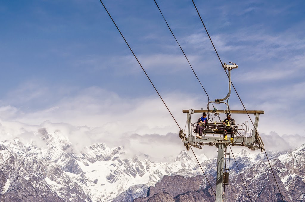 A snowy landscape in Auli, Uttarakhand, featuring ski slopes, a ropeway tower, and majestic snow-capped Himalayan peaks in the background.