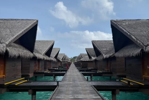 An exterior shot of a long wooden jetty leading to a row of thatched-roof overwater bungalows in a tropical resort, with the clear turquoise water of the ocean.