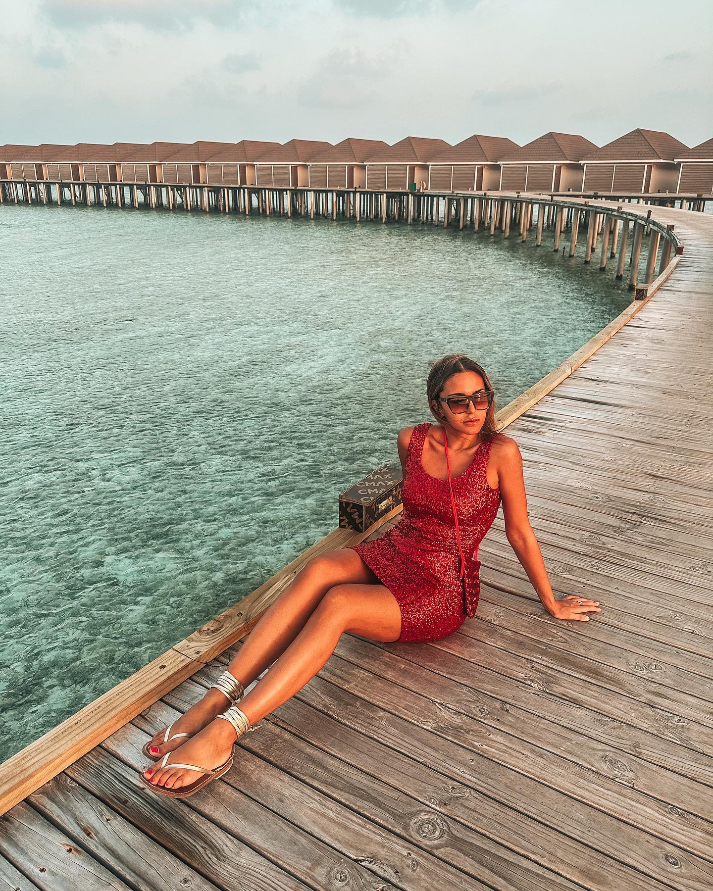 An exterior shot of a long wooden jetty leading to a row of thatched-roof overwater bungalows in a tropical resort, with the clear turquoise water of the ocean.