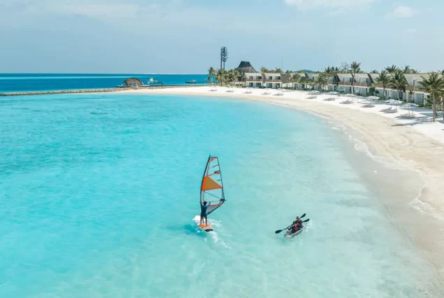 An exterior shot of a large, modern resort in the Maldives, with overwater bungalows and a long water slide leading to a pool, surrounded by the clear turquoise ocean.