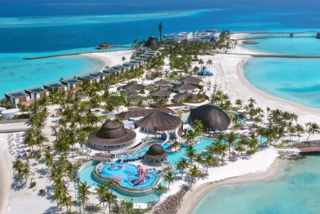 An exterior shot of a large, modern resort in the Maldives, with overwater bungalows and a long water slide leading to a pool, surrounded by the clear turquoise ocean.