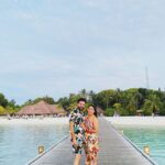 An exterior shot of a long wooden jetty leading to a row of thatched-roof overwater bungalows in a tropical resort, with the clear turquoise water of the ocean.