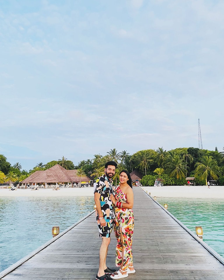 An exterior shot of a long wooden jetty leading to a row of thatched-roof overwater bungalows in a tropical resort, with the clear turquoise water of the ocean.