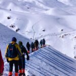 A group of trekkers walking on a snow-covered trail towards the Kuari Pass in the Himalayas, with vast snowfields and a clear sky in the background.