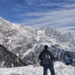 A scenic photograph of a line of trekkers hiking on a rocky mountain path towards Triund Top, with a massive snow-capped Himalayan range dominating the background.