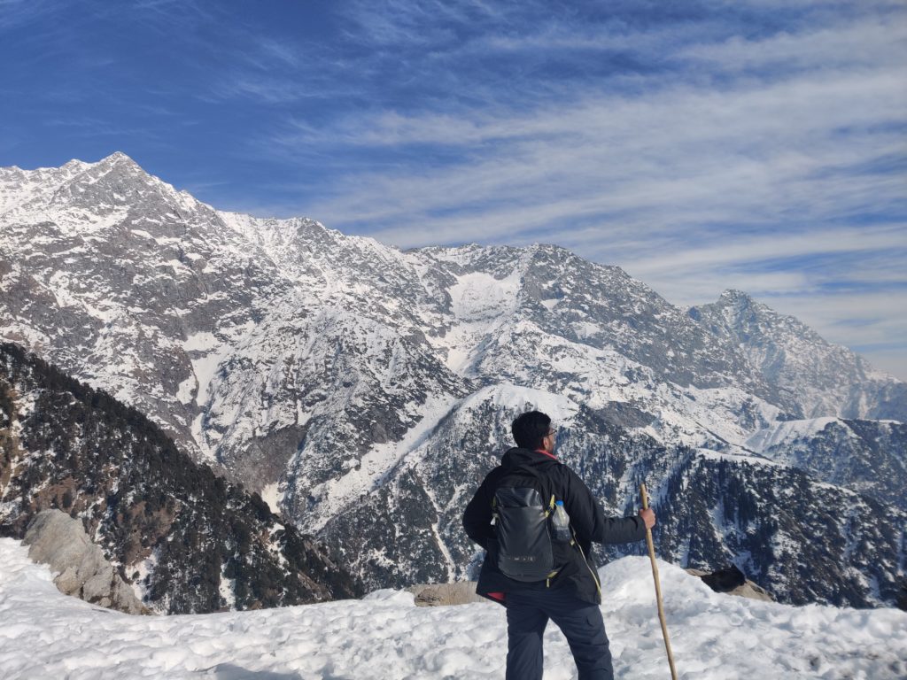 A scenic photograph of a line of trekkers hiking on a rocky mountain path towards Triund Top, with a massive snow-capped Himalayan range dominating the background.
