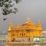 A stunning night view of the Golden Temple in Amritsar, brightly lit and reflected in the sacred Sarovar (pool), with devotees visible around the periphery.