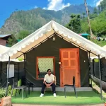 A scenic photograph of a camping site in Kasol, with several colorful tents pitched on the banks of a river, surrounded by pine trees and mountains.
