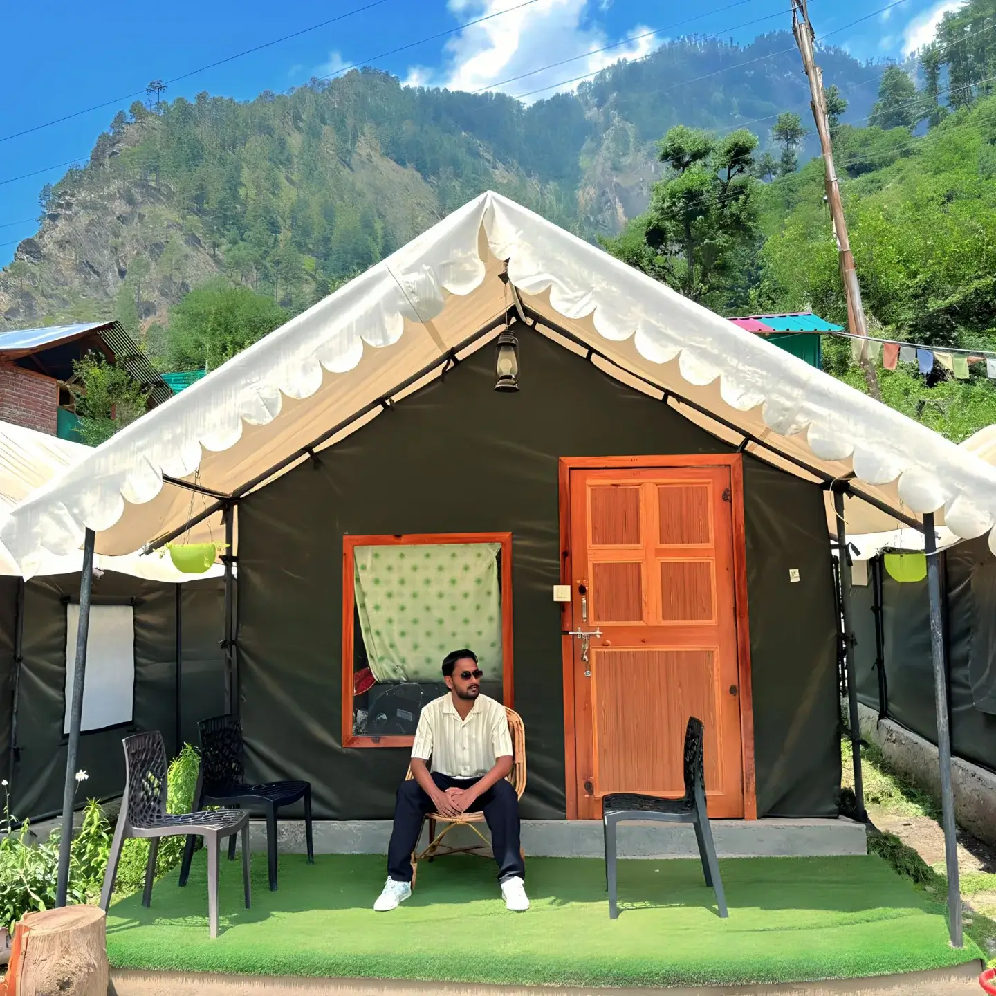 A scenic photograph of a camping site in Kasol, with several colorful tents pitched on the banks of a river, surrounded by pine trees and mountains.