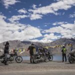 A high-altitude road cutting through a vast, barren desert landscape in Ladakh, with towering mountains and a car in the distance, emblematic of a road trip to Leh-Ladakh.