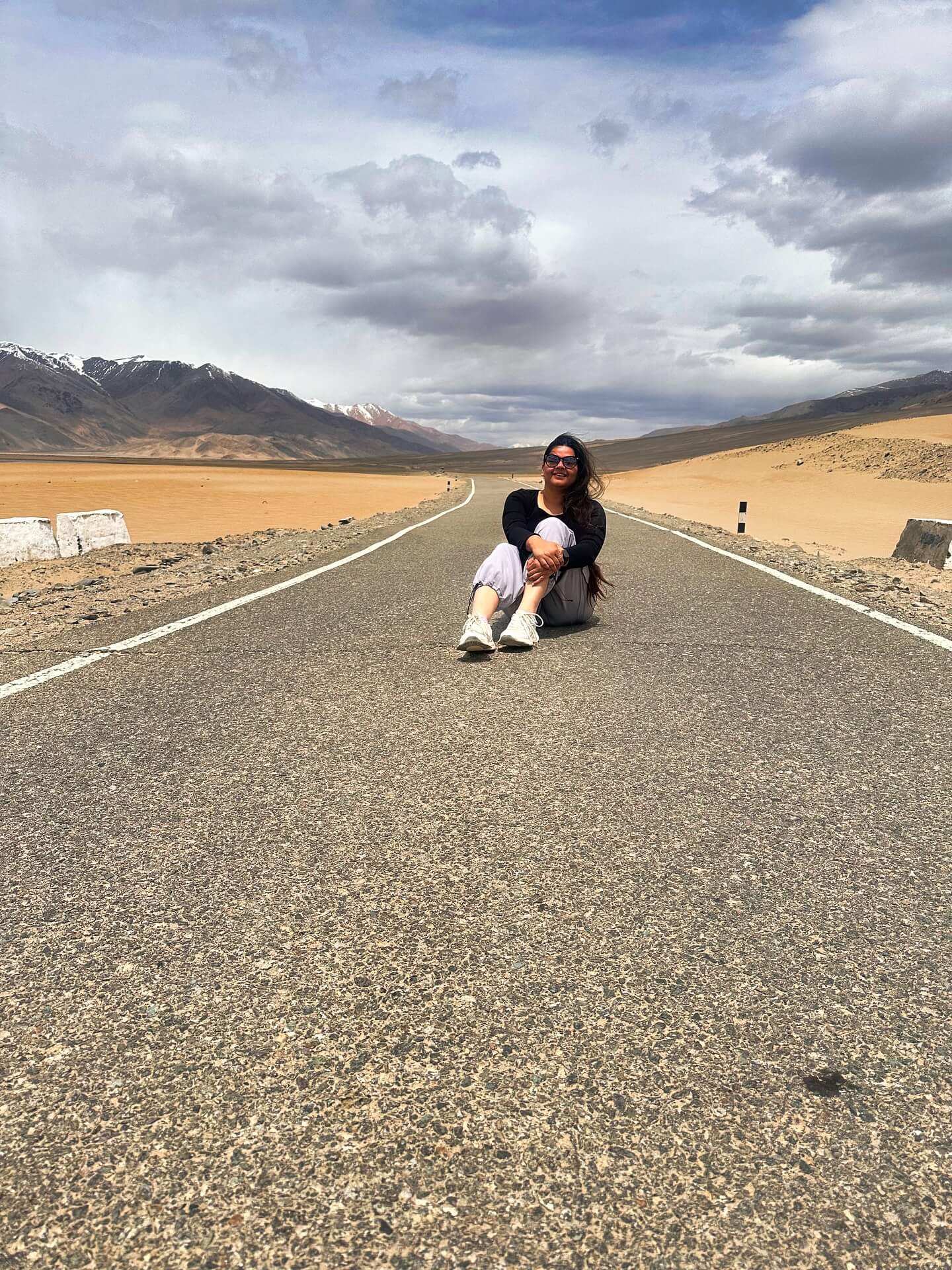 A car driving on a long, winding road through the high-altitude, barren landscape of Ladakh, with snow-capped mountains in the background.