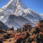 A high-altitude mountain landscape on the Manaslu Circuit Trek, showing a winding, snow-covered trail and prayer flags against a backdrop of massive, jagged Himalayan peaks.