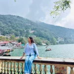 A high-angle, panoramic view of the crescent-shaped Naini Lake surrounded by hills dotted with buildings, showcasing Nainital: The Lake City.
