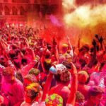 A vibrant, chaotic scene of people celebrating Holi in Pushkar, covered head-to-toe in bright, dry colored powder (gulal) while dancing and cheering.