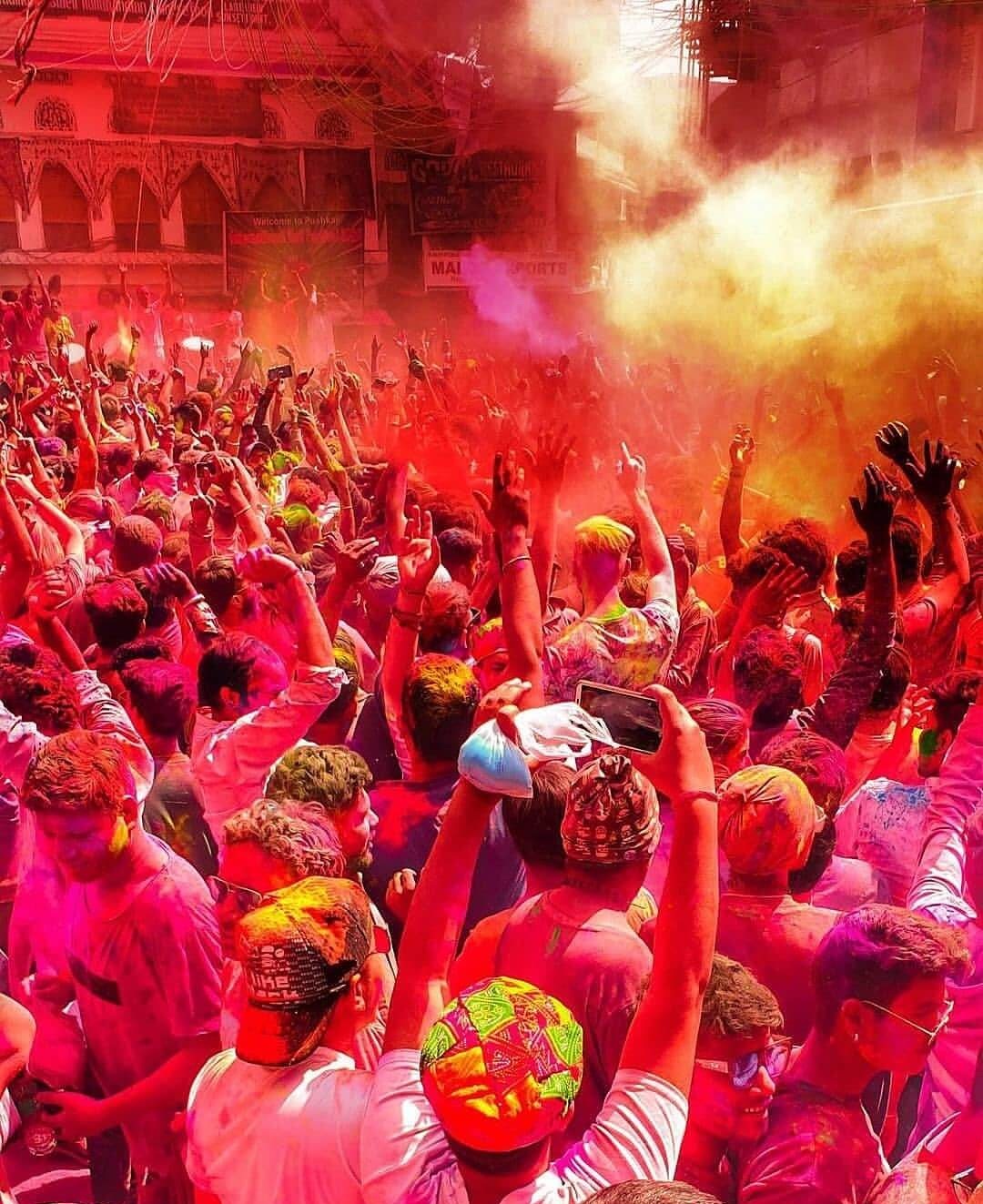 A vibrant, chaotic scene of people celebrating Holi in Pushkar, covered head-to-toe in bright, dry colored powder (gulal) while dancing and cheering.