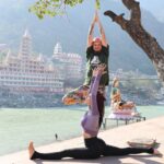 A person performing a yoga pose in a serene, natural setting with mountains and a river in the background.