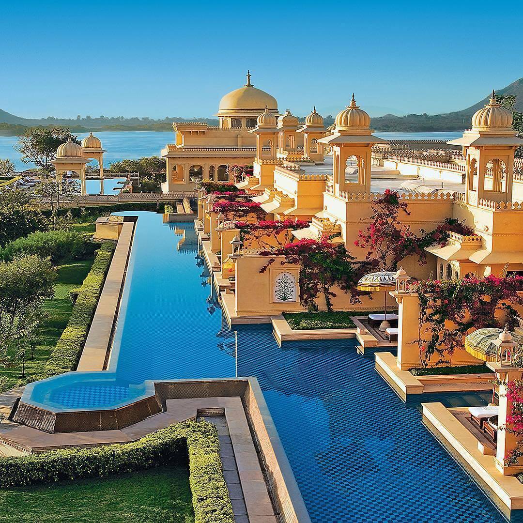 A magnificent view of The Oberoi Udaivilas palace resort on the bank of Lake Pichola in Udaipur, featuring ornate domes and interconnected courtyards.