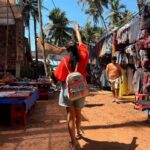 A colorful, crowded scene at the Anjuna Flea Market in Goa, showing stalls packed with textiles, jewelry, and handicrafts, with many shoppers browsing.
