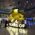 An interior shot of the Hamad International Airport terminal in Doha, showing its modern design with a high, arched ceiling, numerous shops and restaurants, and the iconic "Lamp Bear" statue in the center.