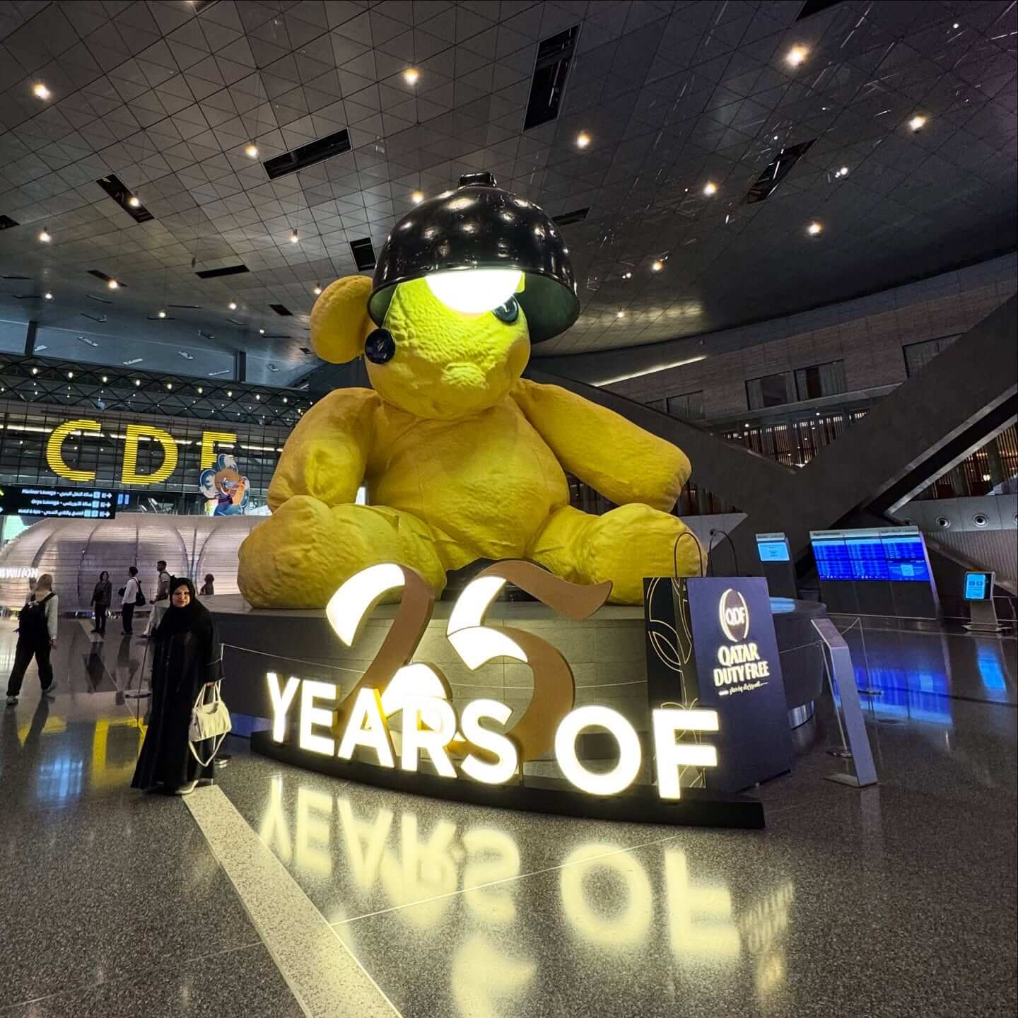 An interior shot of the Hamad International Airport terminal in Doha, showing its modern design with a high, arched ceiling, numerous shops and restaurants, and the iconic "Lamp Bear" statue in the center.