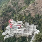 An exterior shot of the Vaishno Devi shrine, showing the temple complex nestled on a mountain, with a large crowd of devotees and the surrounding hills in the background.