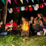 A vibrant image of a campfire under a starry night sky with tents pitched in the background