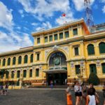 An exterior shot of the Central Post Office in Ho Chi Minh City, a yellow building with green trim, arched windows, and a large clock above the main entrance.