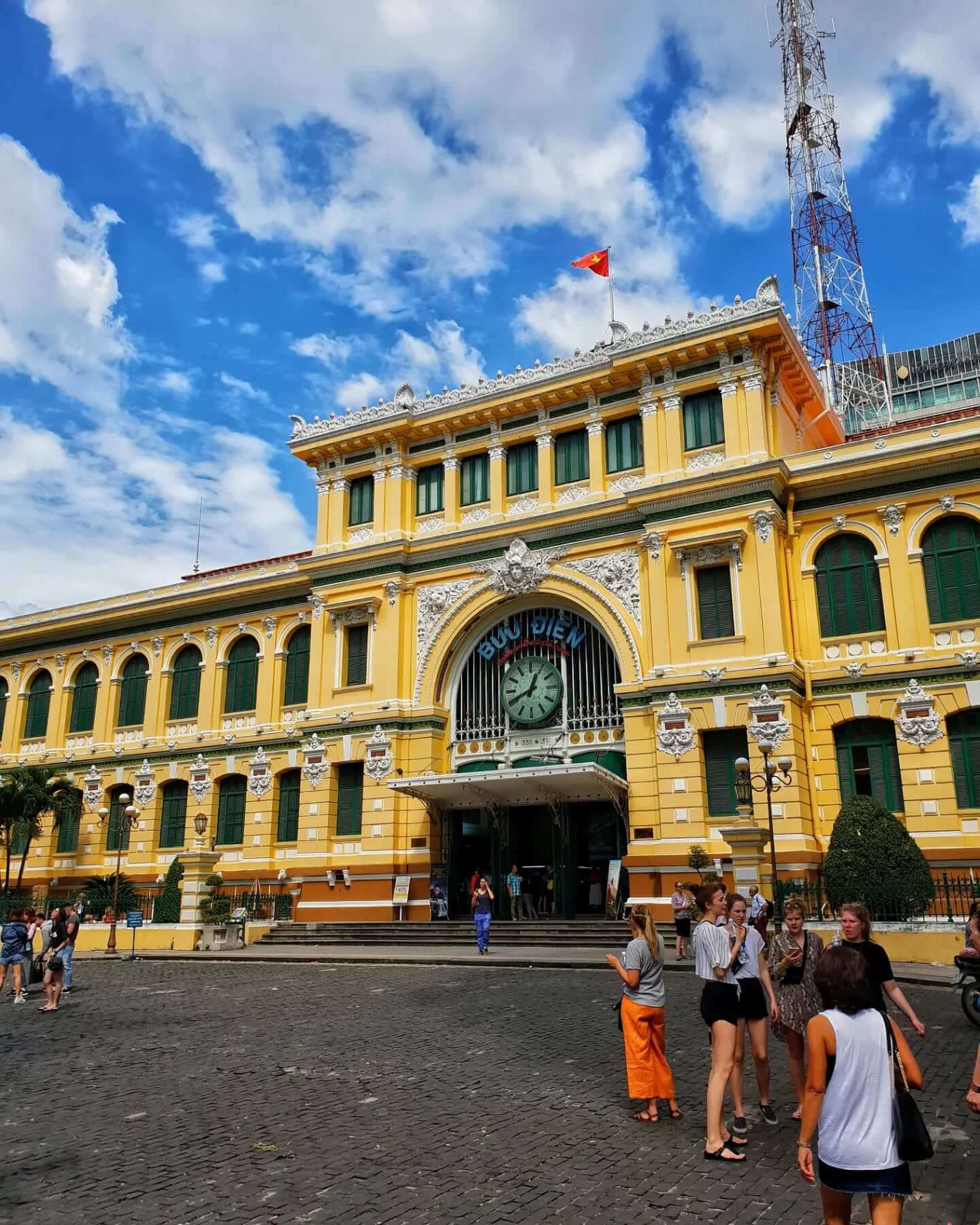 An exterior shot of the Central Post Office in Ho Chi Minh City, a yellow building with green trim, arched windows, and a large clock above the main entrance.