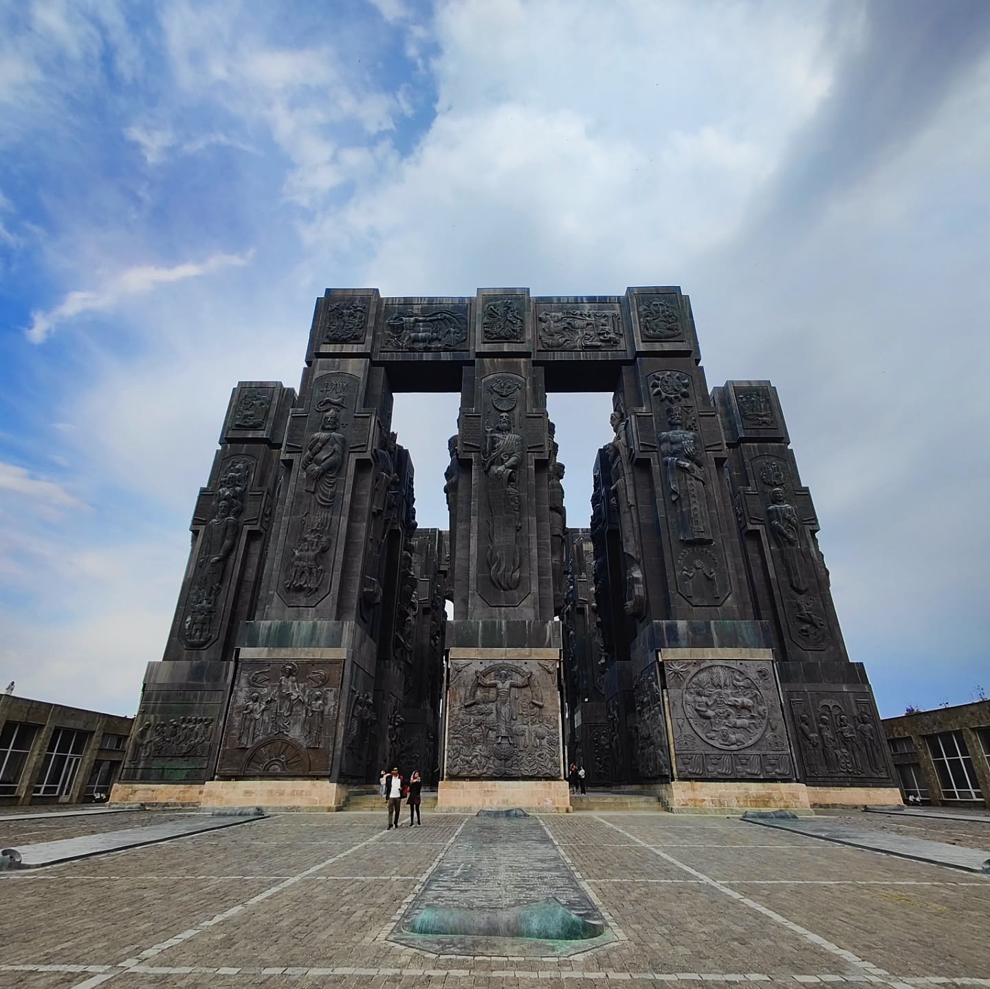 An exterior shot of the Chronicles of Georgia monument, a massive stone sculpture with tall columns depicting kings, queens, and religious figures, located on a hill overlooking Tbilisi.