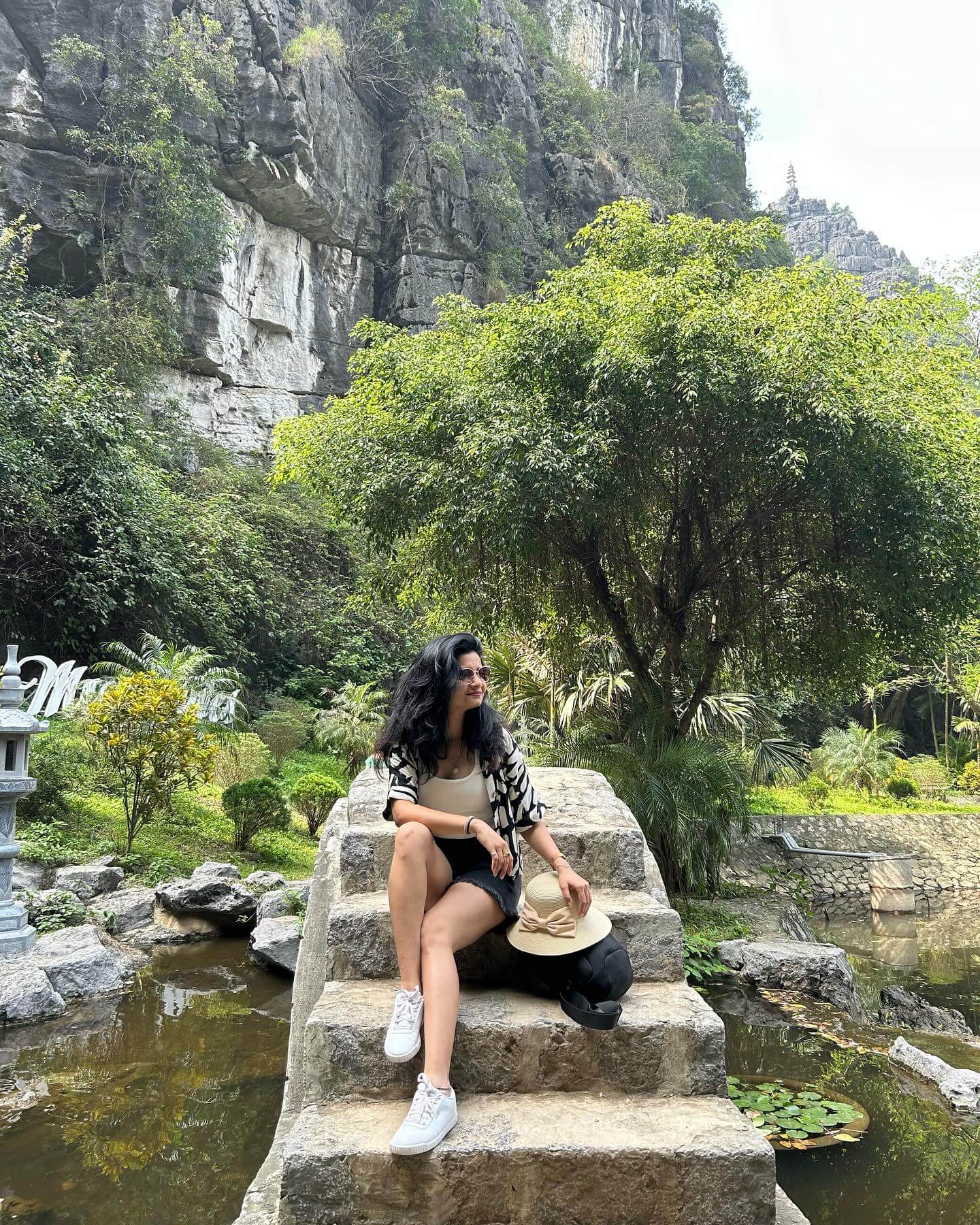 An exterior shot of a person on a traditional wooden boat navigating a serene river surrounded by lush green rice paddies and towering limestone mountains.