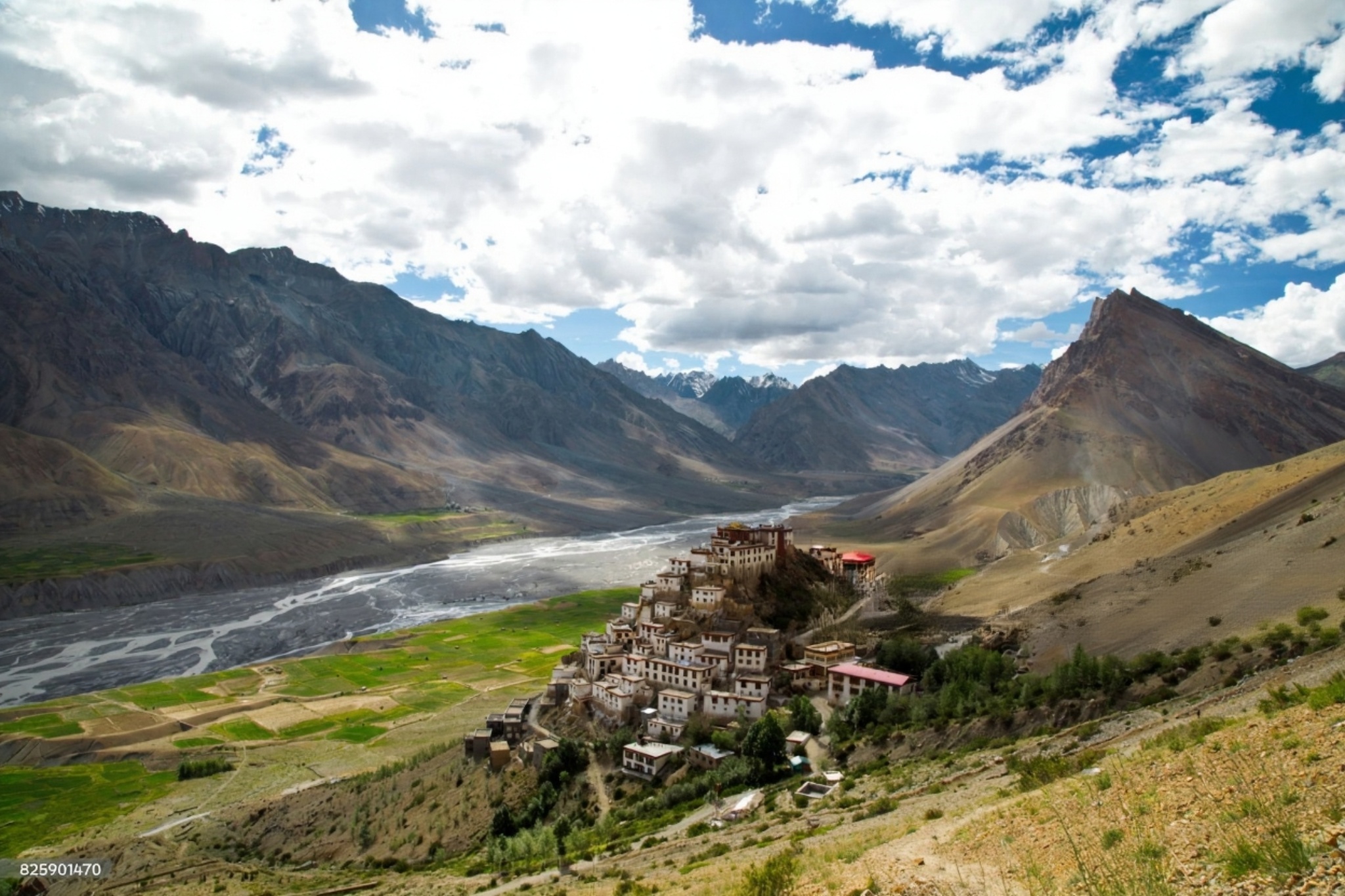 A panoramic view of the vast, rugged landscape of Spiti Valley, showing barren, brown mountains with a winding road, and a river flowing through the valley under a clear blue sky.