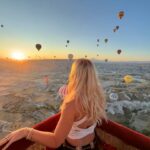 A panoramic photograph of a sky filled with dozens of colorful hot air balloons floating over a hilly landscape at sunrise, with the sun's warm light illuminating the scene.