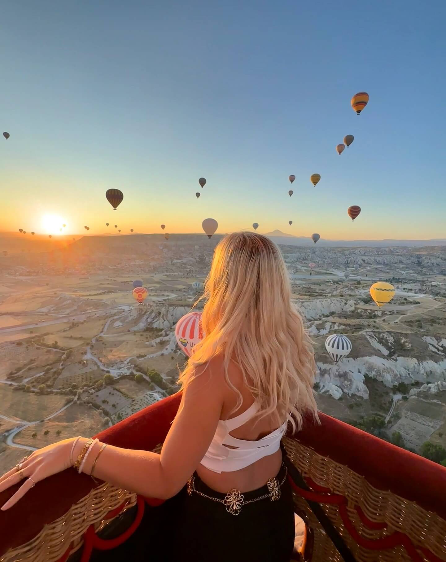 A panoramic photograph of a sky filled with dozens of colorful hot air balloons floating over a hilly landscape at sunrise, with the sun's warm light illuminating the scene.