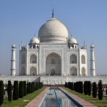 An exterior shot of the Taj Mahal in Agra, India, a large white marble mausoleum with a central dome, surrounded by symmetrical minarets and reflected in a long pool of water.