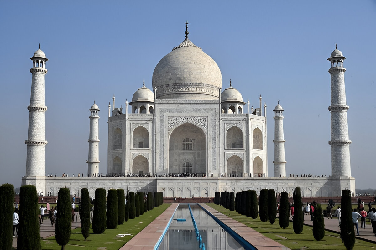 An exterior shot of the Taj Mahal in Agra, India, a large white marble mausoleum with a central dome, surrounded by symmetrical minarets and reflected in a long pool of water.