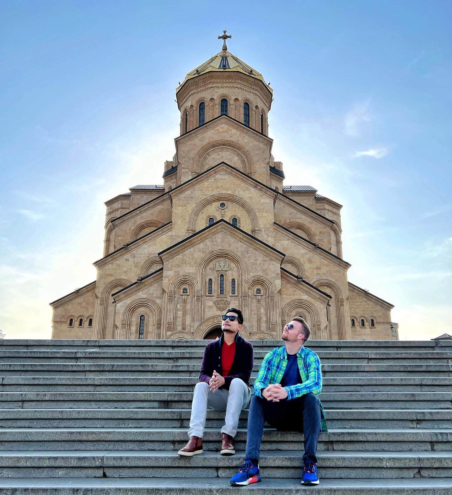An exterior shot of the Tsminda Sameba Cathedral in Tbilisi, showing a large, modern Georgian Orthodox church with its golden-domed main cupola and a bell tower against a blue sky.