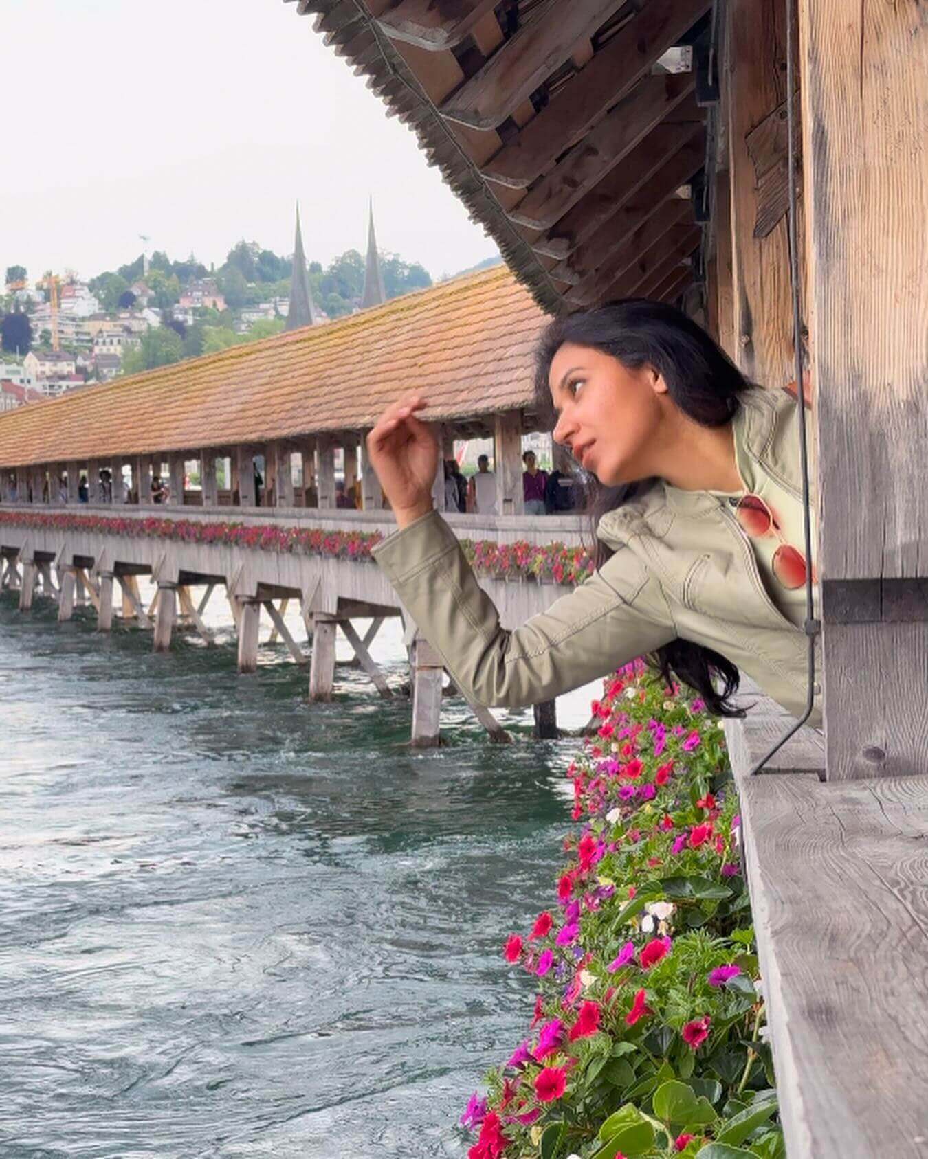 An exterior shot of the Chapel Bridge (Kapellbrücke) in Lucerne, a historic wooden footbridge with a distinctive roof, a prominent water tower, and a view of the city and a clear river.
