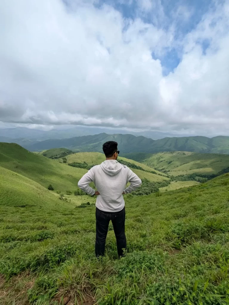 A panoramic view of a lush green landscape in Chikmagalur, with rolling hills covered in dense vegetation and coffee plantations under a cloudy sky.