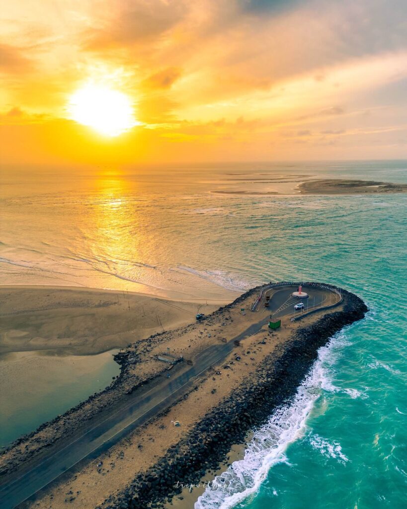 A desolate, sandy beach landscape in Dhanushkodi, with the ruins of a stone building in the distance and a calm, turquoise sea stretching out under a clear sky.