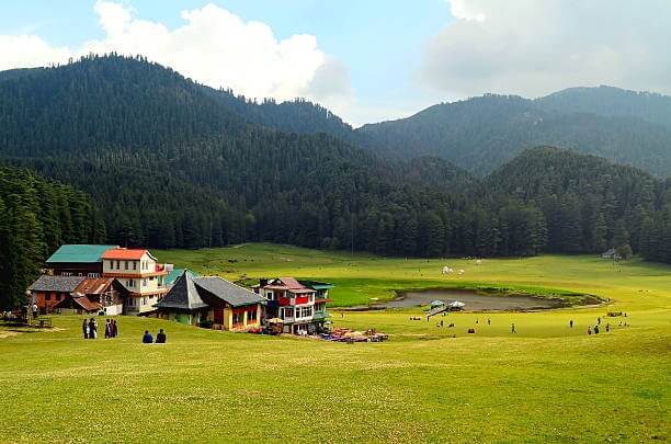 A scenic view of Khajjiar's green meadow with a lake at its center, set against a backdrop of snow-capped mountains in Himachal Pradesh.