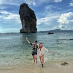A pristine white sandy beach on Koh Poda Island in Krabi, Thailand, with clear turquoise water, a few longtail boats, and a distinctive limestone rock formation just offshore.