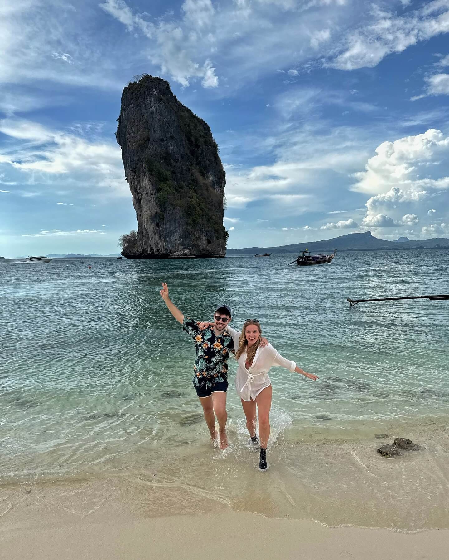 A pristine white sandy beach on Koh Poda Island in Krabi, Thailand, with clear turquoise water, a few longtail boats, and a distinctive limestone rock formation just offshore.