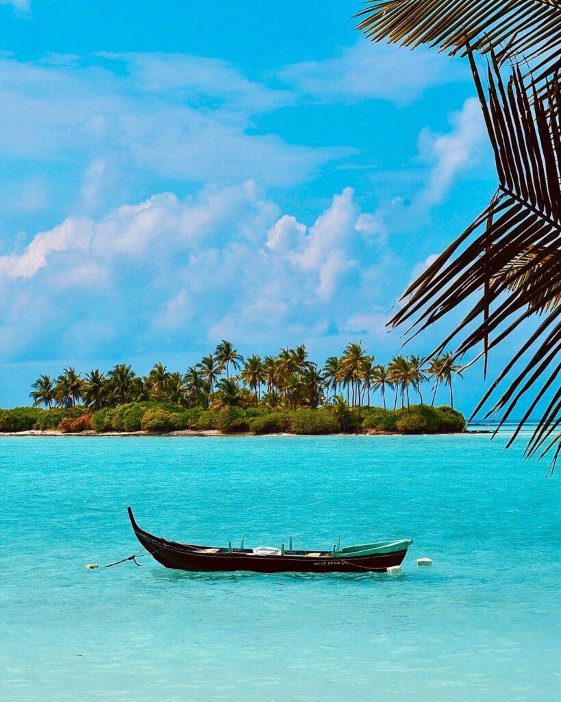 An aerial photograph of a small tropical island in Lakshadweep, with a thin strip of white sand surrounding lush green vegetation, bordered by clear turquoise water and a coral reef.