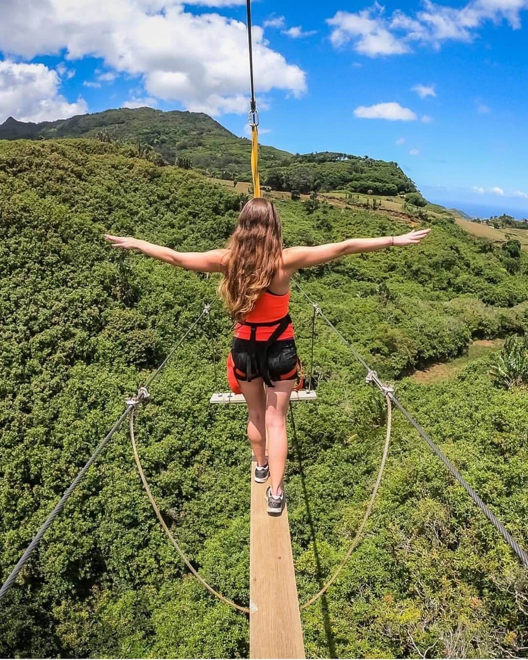 An exterior shot of the Nepalese Bridge in Switzerland, a long, narrow wooden suspension bridge crossing a deep gorge with a view of a waterfall and the surrounding lush green forest.