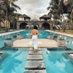 An exterior shot of the Outrigger Mauritius Beach Resort, showing the large swimming pool with palm trees, lounge chairs, and a view of the ocean.
