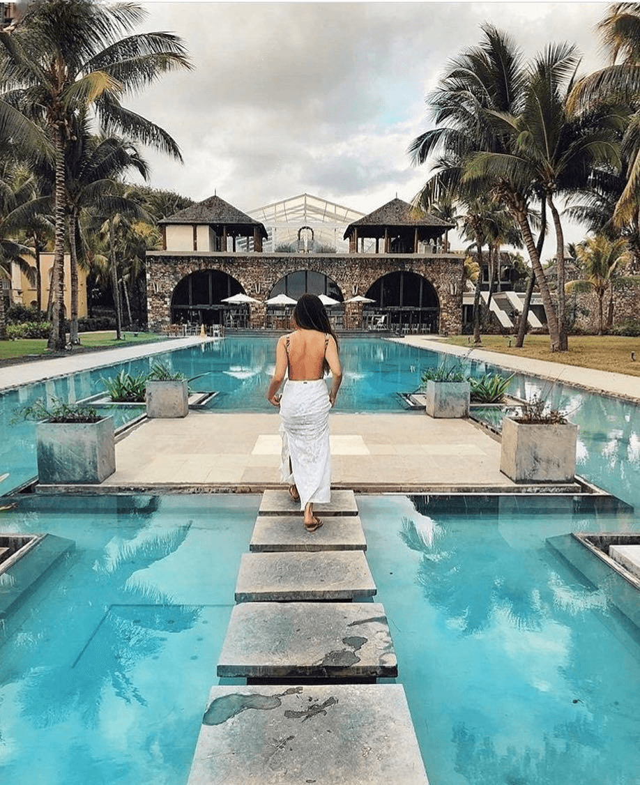 An exterior shot of the Outrigger Mauritius Beach Resort, showing the large swimming pool with palm trees, lounge chairs, and a view of the ocean.