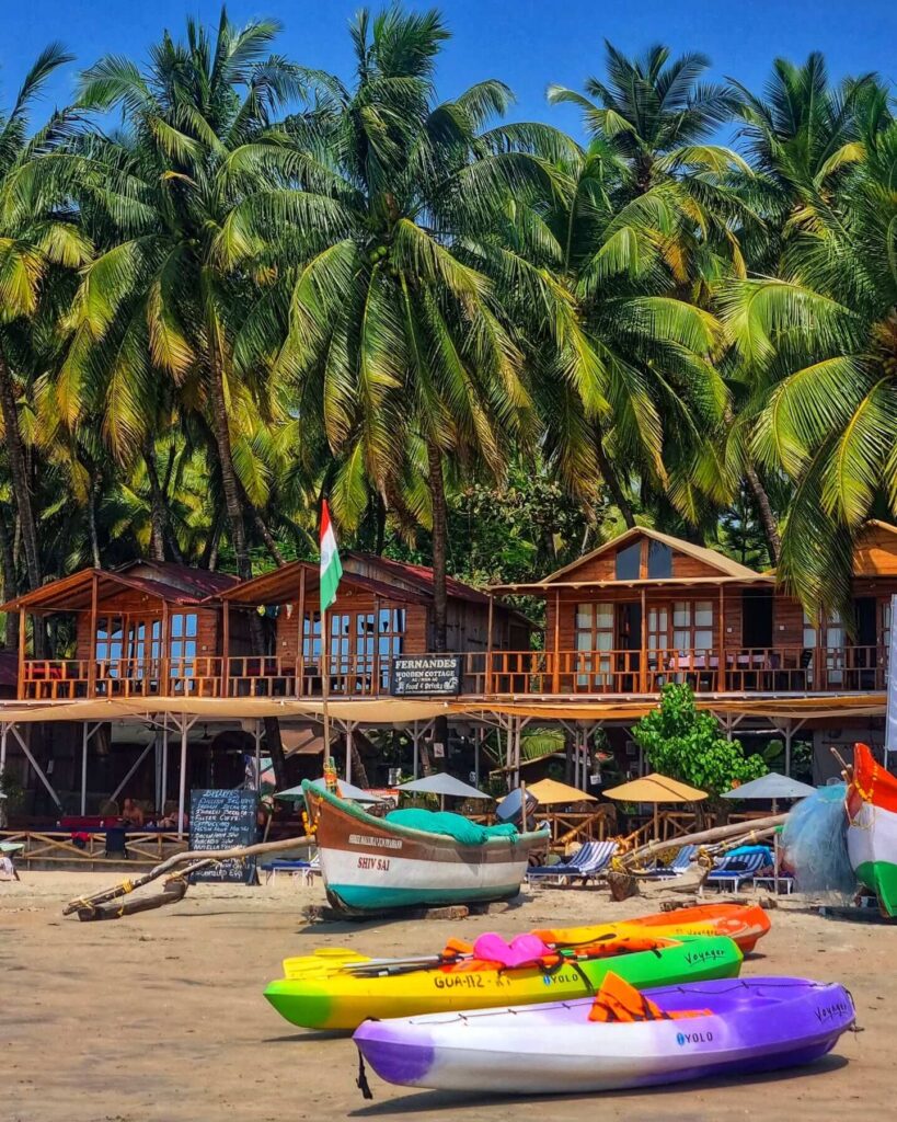 A serene photograph of Palolem Beach in Goa during sunset, showing a crescent-shaped bay with calm water and a few small boats near the shore, surrounded by palm trees and lush green hills, with the sky displaying a mix of warm orange, pink, and purple colors.