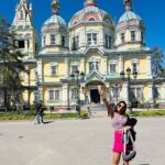 A photograph of the Zenkov Cathedral, a bright yellow and green Russian Orthodox cathedral made of wood, set against a backdrop of trees in a park.