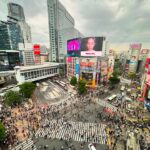 An aerial view of the Shibuya Crossing in Tokyo, showing a massive crowd of pedestrians crossing from multiple directions, surrounded by tall buildings adorned with vibrant neon signs and digital billboards at dusk.